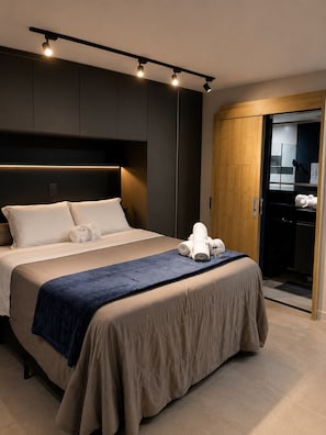 A modern hotel room with a large bed, a wooden wardrobe, and a bathroom visible through an open door. - Moderno e Equipado | 1 Vaga | Metrô República (São Paulo)