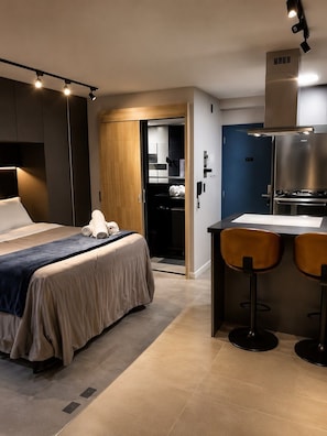 Ein modernes Hotelzimmer mit einem Bett, einer kleinen Küchenzeile und einem durchgehenden Türspalt sichtbaren Badezimmer.