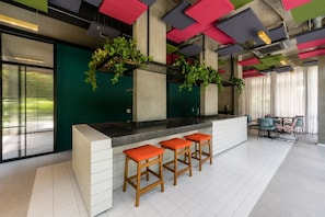 A modern bar area with a white counter, three wooden stools with orange cushions, and a green wall. - Vista | Centro SP | Metrô República (Santa Ifigênia)