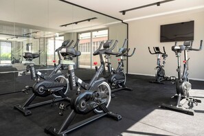 A gym with stationary bikes, a large mirror, and a TV on the wall. - 1 Dormitório | 1 Vaga no coração do Brooklin (São Paulo)