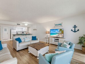 A living room with a white sofa, a coffee table, and a TV displaying a beach scene. There is a kitchen in the background with white cabinets and a sign that says 'Welcome to the beach.' - Sandy Shores #202 (Madeira Beach)