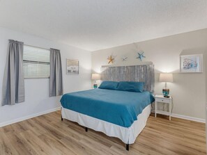 A bedroom with a bed, curtains, a headboard, starfish decorations, a map, and a nightstand with a lamp. - Sandy Shores #202 (Madeira Beach)