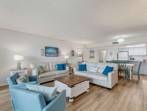 A modern living room with a sofa, chairs, a coffee table, and a dining area with a kitchen in the background. - Sandy Shores #202 (Madeira Beach)