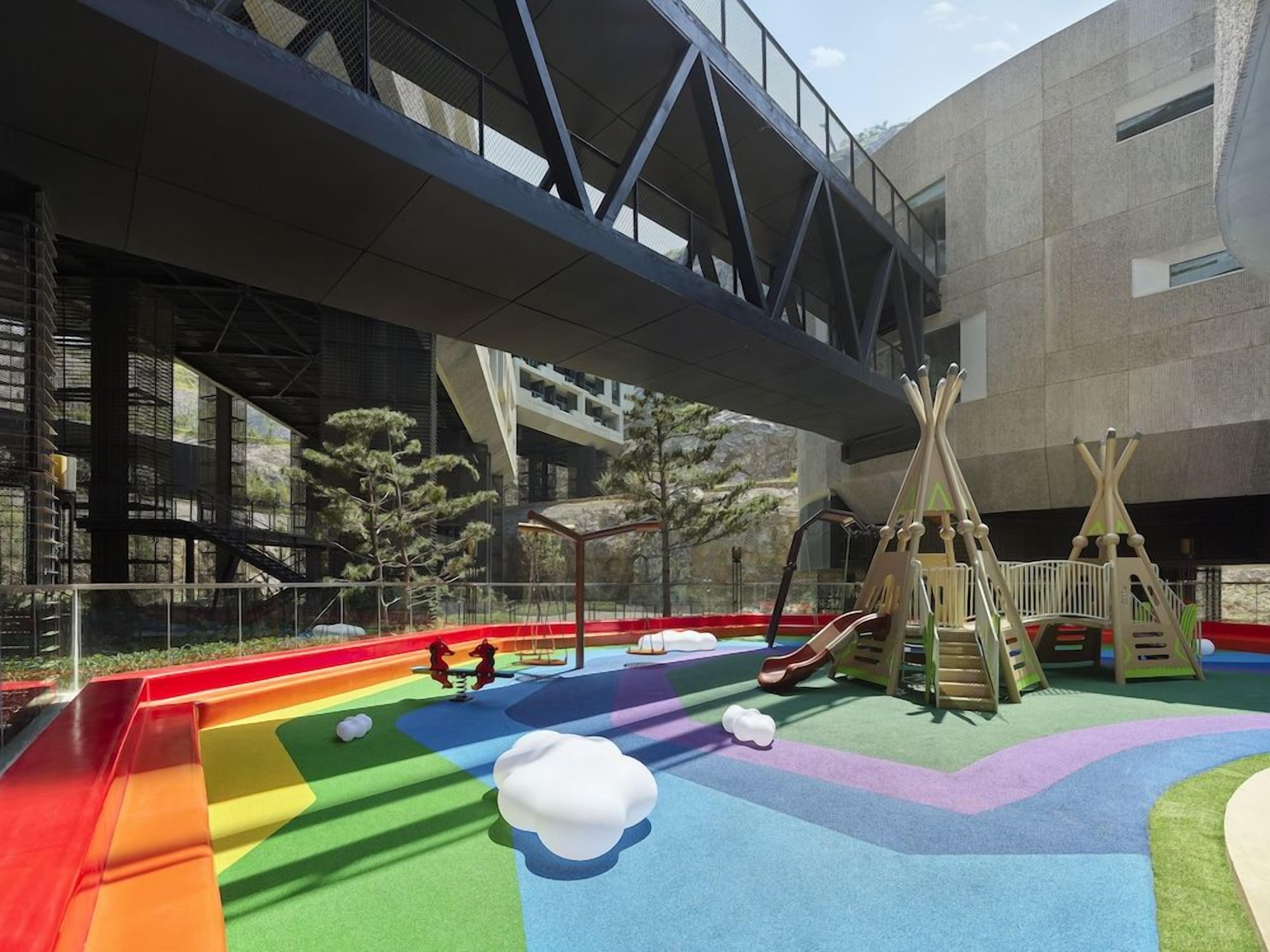 An outdoor playground with slides and climbing structures, set against a modern building with a bridge overhead.