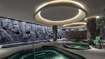 A modern indoor swimming pool area with multiple pools, a rock wall, and a unique ceiling design.