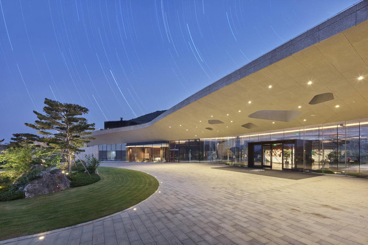 A modern building with a curved roof and glass facade, a paved entrance area, and a landscaped garden with a tree and rocks.