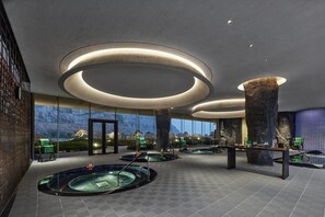 Un moderno espacio de spa con varias jacuzzis, una gran piedra central y vistas a montañas a través de amplios ventanales.