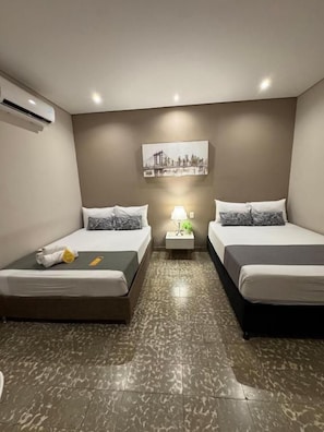 Habitación de hotel con dos camas, una mesita de noche con lámpara y aire acondicionado de pared.