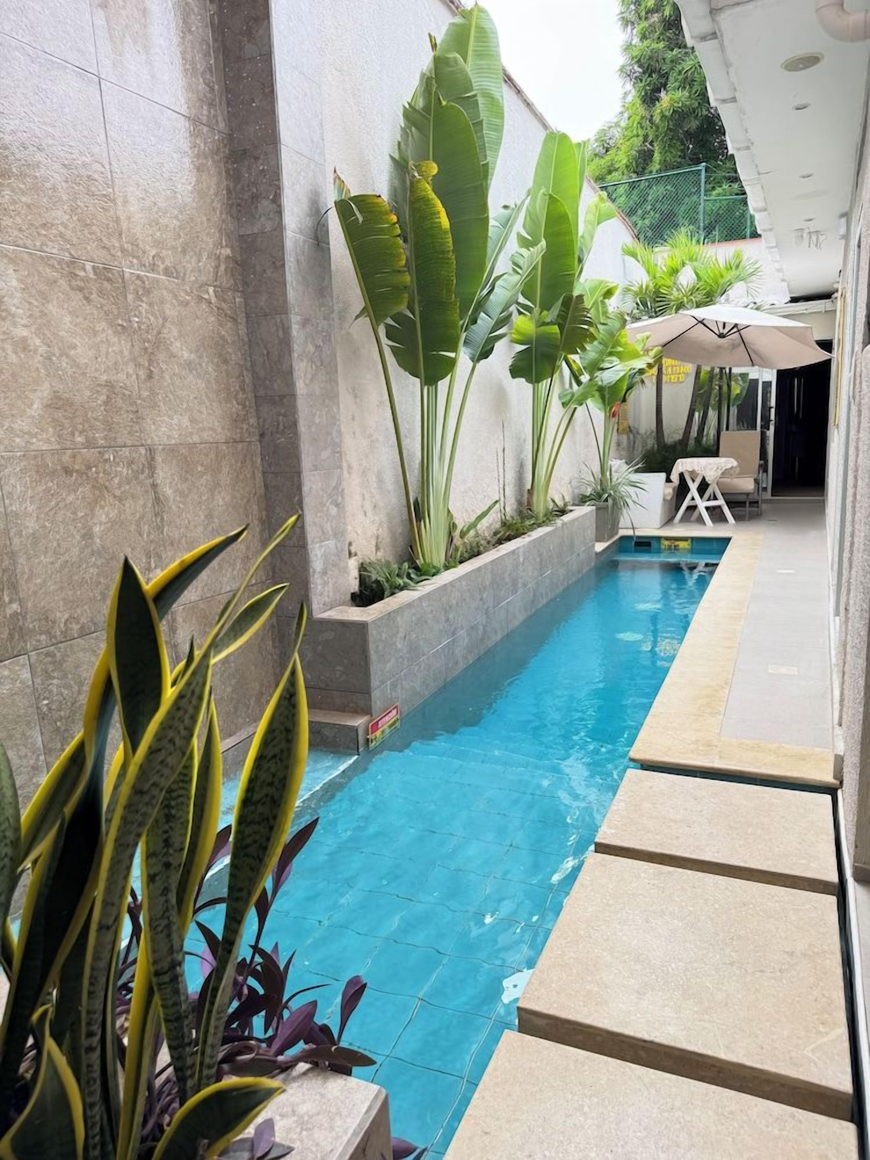 Une piscine dont le bord est revêtu de carreaux, entourée de plantes et dotée d’un espace détente abrité.