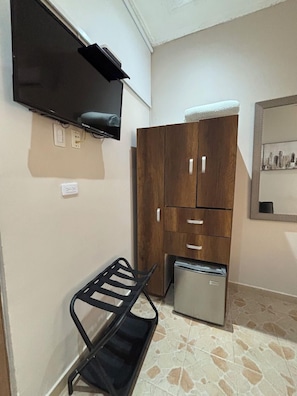 Una habitación con una televisión de pantalla plana montada en la pared, un armario de madera, un refrigerador pequeño, un estante de metal negro y piso de baldosas.
