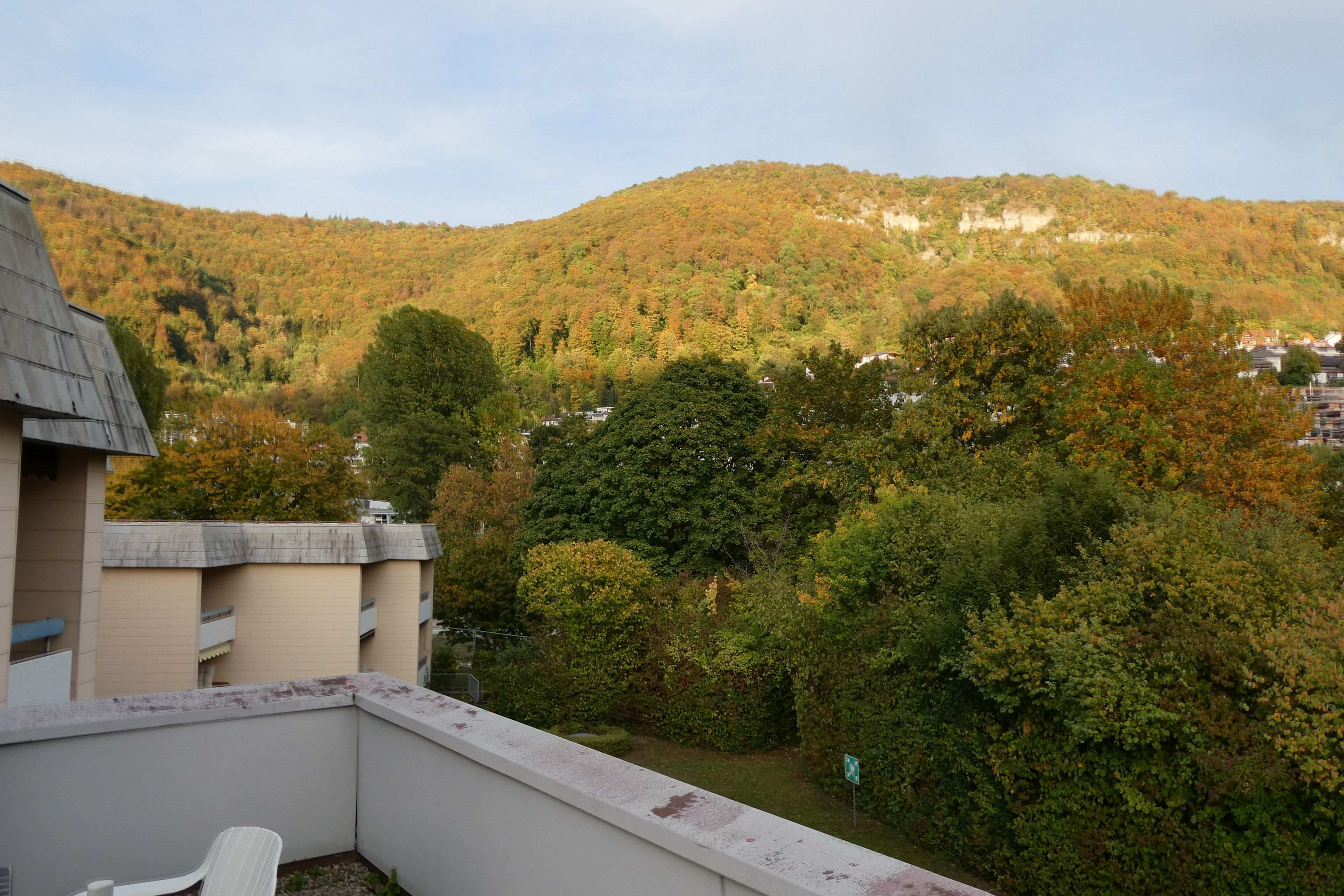 Ein Balkon mit Blick auf einen Hügel, der mit Herbstlaub bedeckt ist, ein Gebäude mit flachem Dach und ein klarer Himmel.