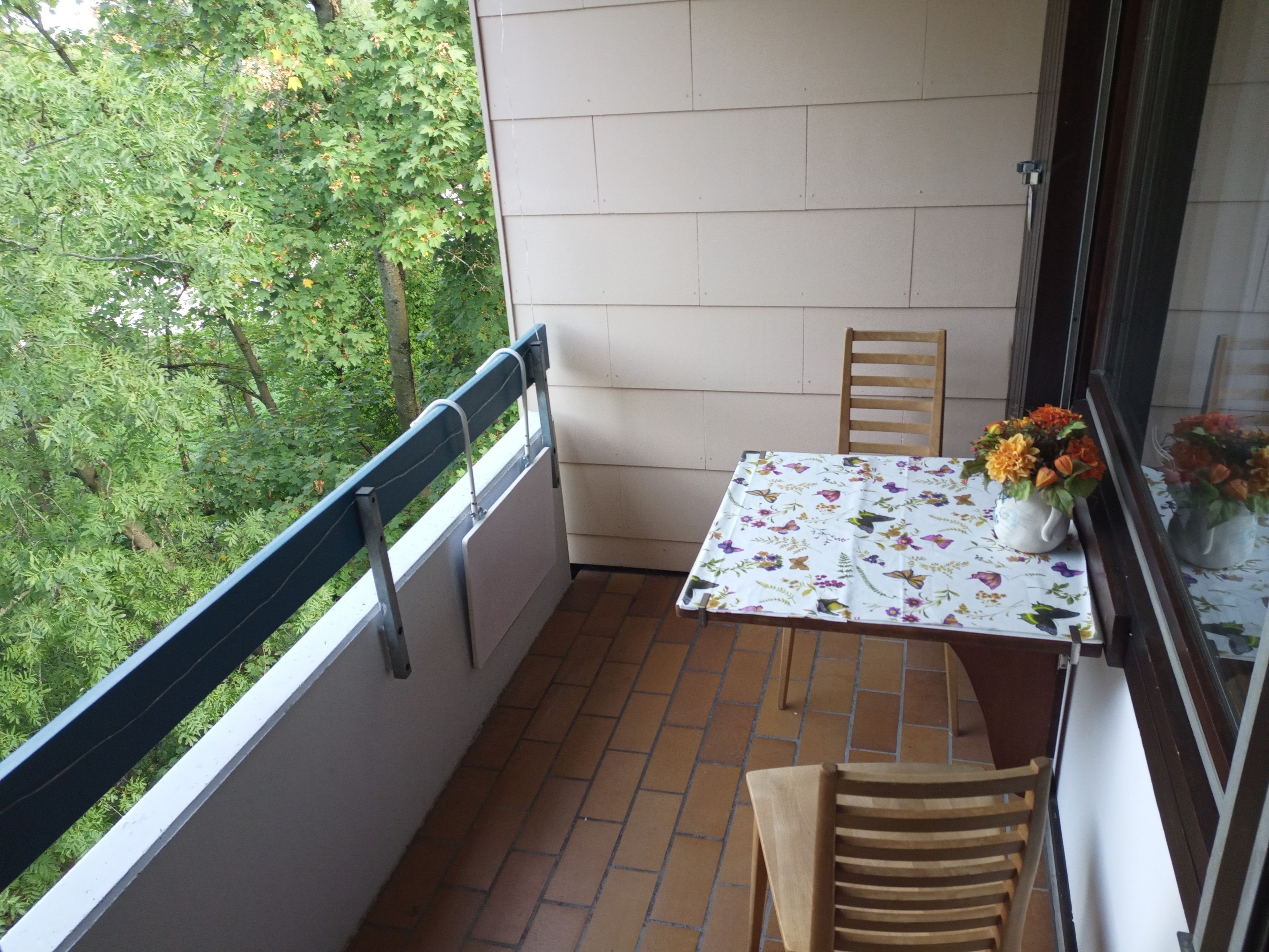 Ein kleiner Balkon mit Tisch, Stühlen und einem Fenster mit Blick auf Grünflächen.
