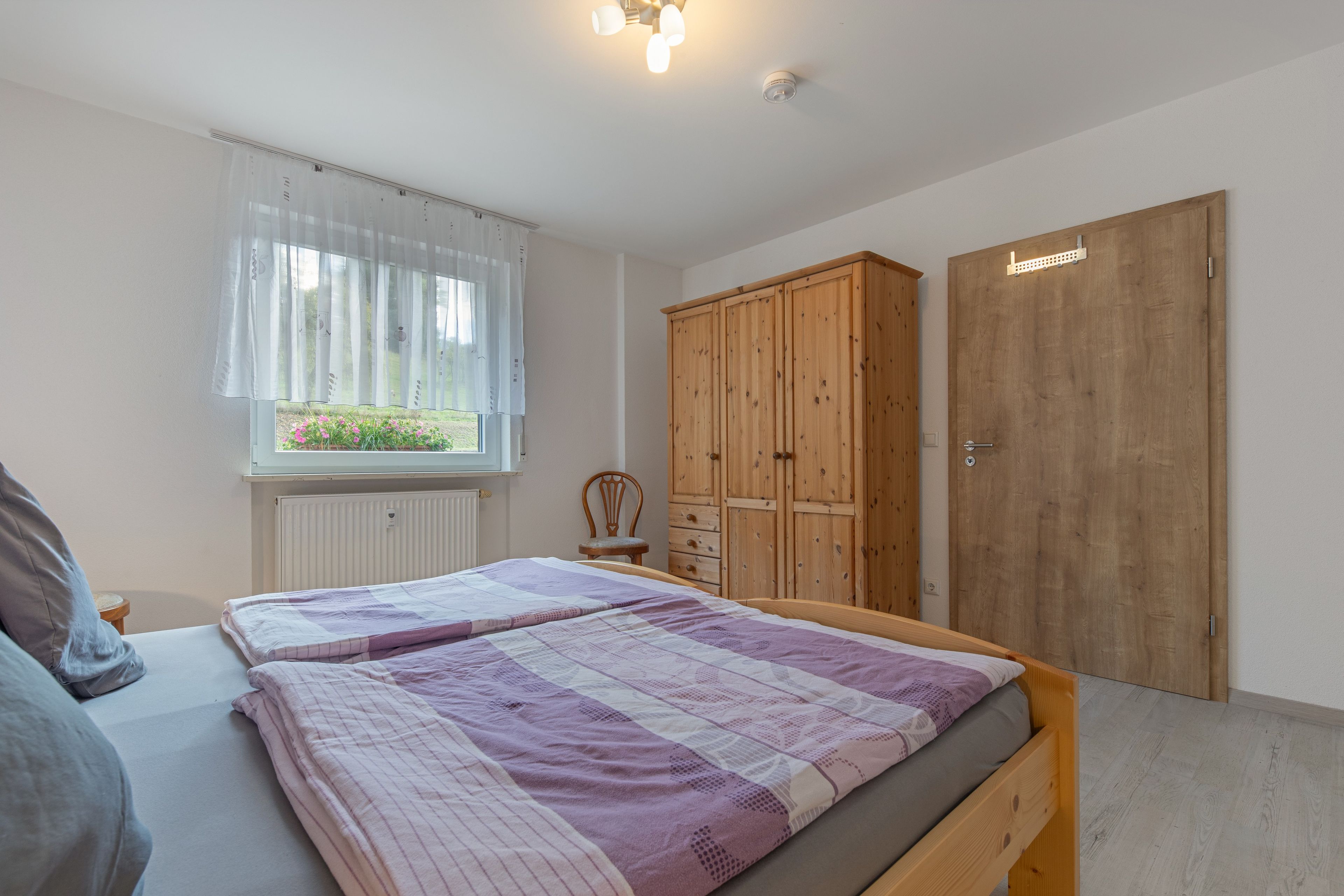 A bedroom with a bed, a wooden wardrobe, a window with curtains, and a radiator.