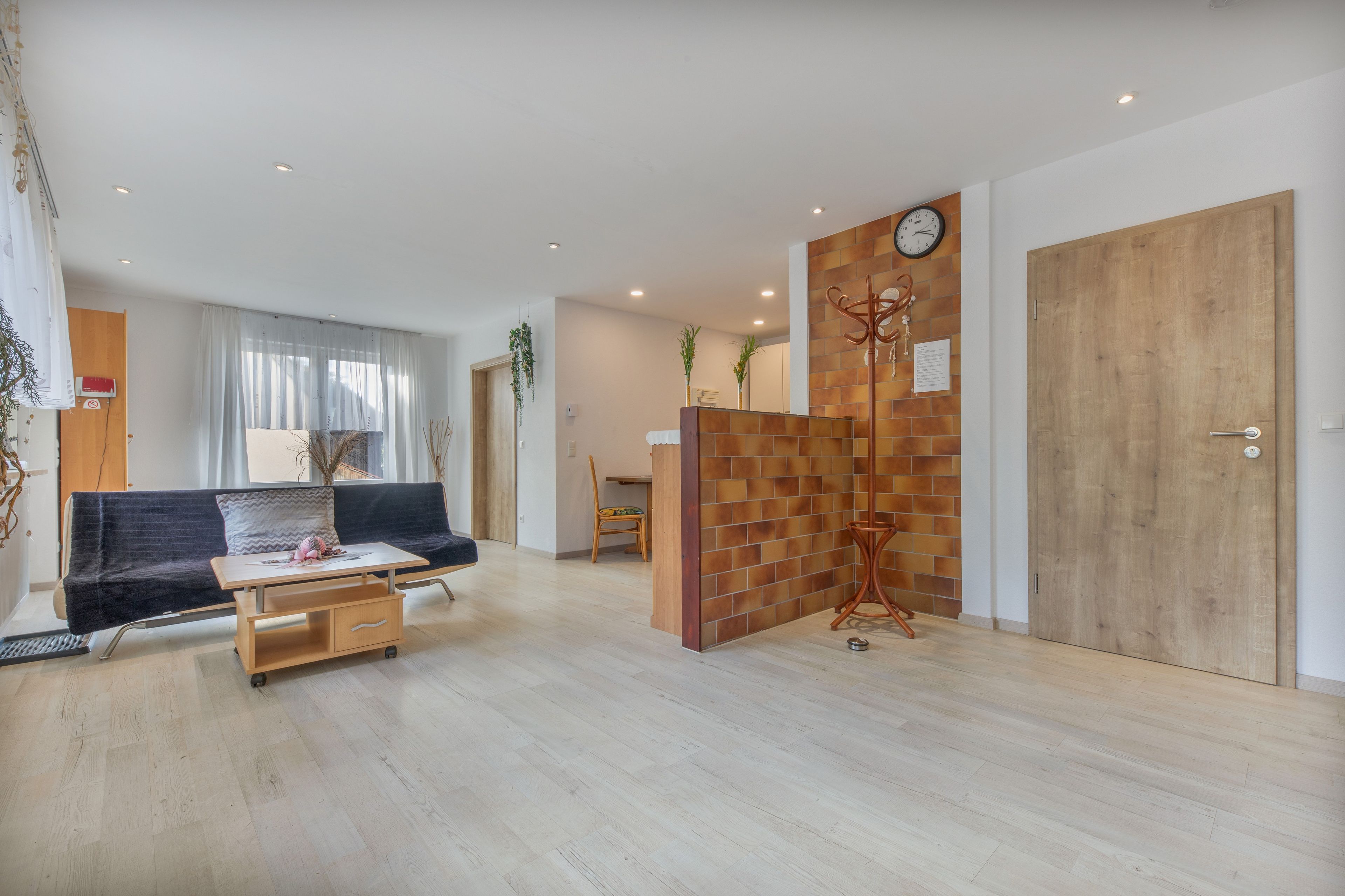 Ein modernes Wohnzimmer mit einer Couch, einem Couchtisch und einer Holzkommode. Eine Holztür und eine Uhr sind an der Wand zu sehen.