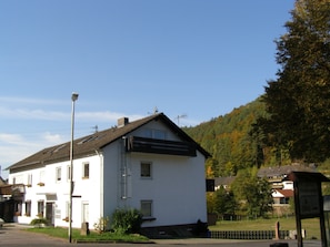 A white building with a sloped roof, a visible balcony, and a small garden area in front.