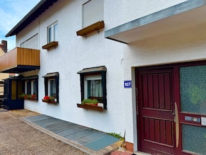 Un bâtiment blanc avec une porte rouge, portant le numéro 107, et des appuis de fenêtre en bois ornés de jardinières.