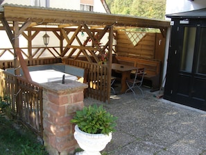 Un espace extérieur aménagé en patio, doté d’une pergola en bois, d’un jardin en briques agrémenté de plantes vertes et d’un coin salon comprenant une table et des chaises.
