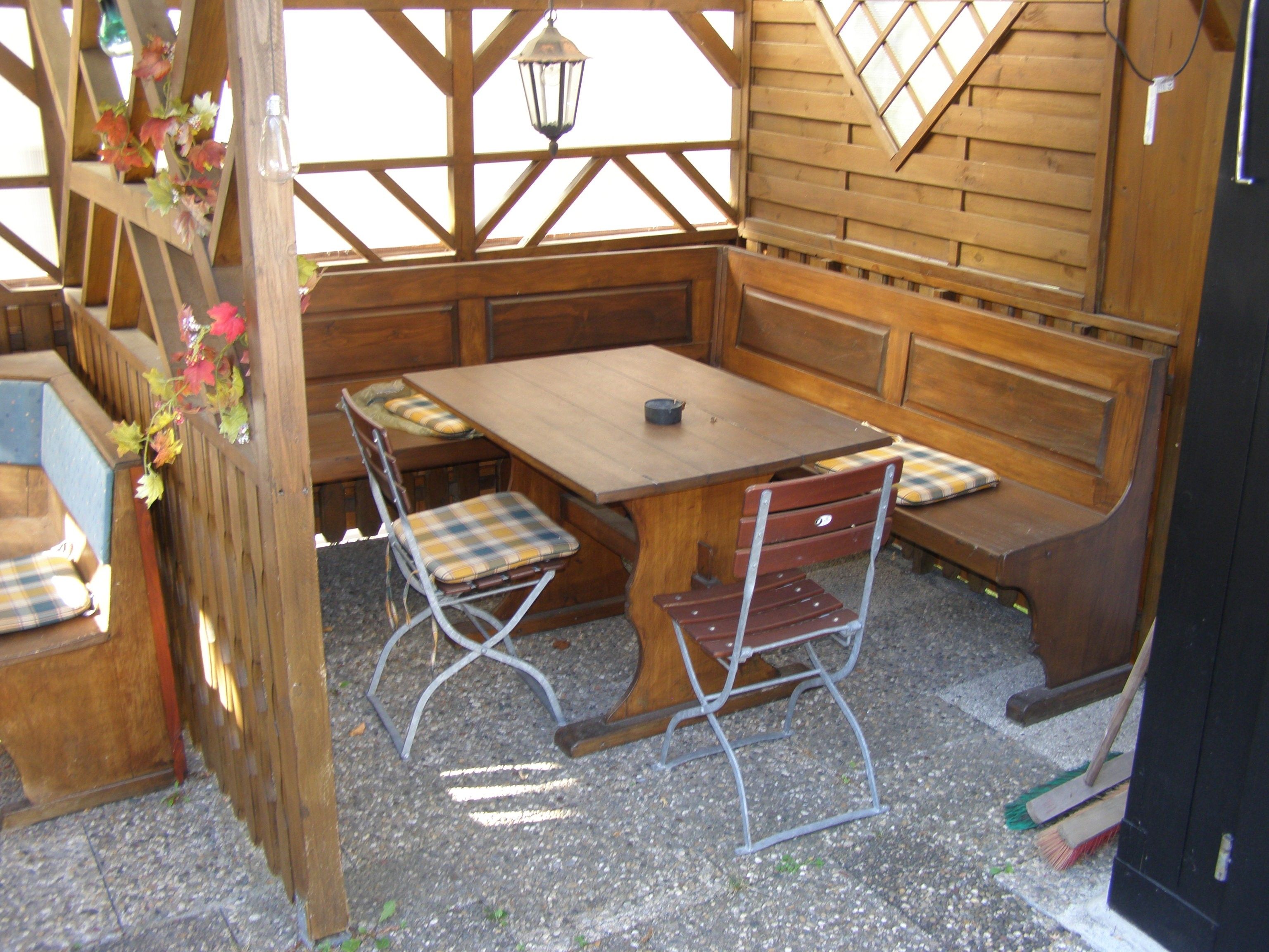 Un área de estar al aire libre con madera, que incluye una mesa y bancos, rodeada por una valla y un muro de madera con ventanas.