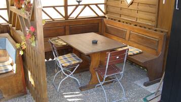 Un área de estar al aire libre con madera, que incluye una mesa y bancos, rodeada por una valla y un muro de madera con ventanas.