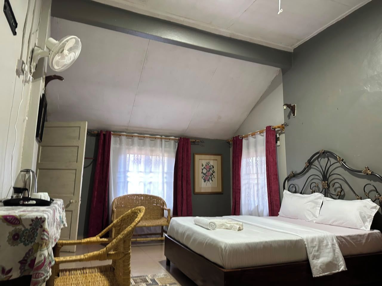 Un dormitorio con una cama, una silla, una mesa con hervidor y una ventana con cortinas.
