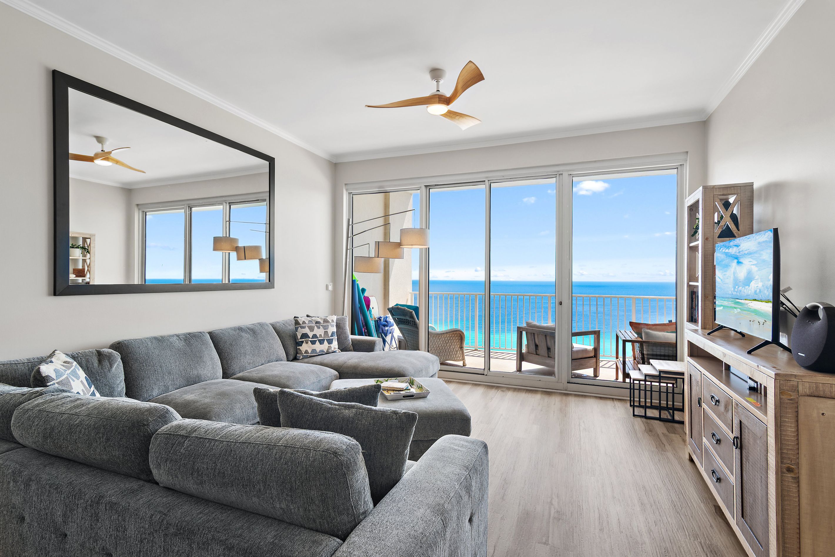 A modern living room with a grey sectional sofa, a ceiling fan, a TV on a wooden cabinet, and a balcony with a view of the ocean.