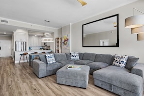 A modern living room with a grey sectional sofa, a coffee table with books, a large mirror, and a kitchen in the background. - Luxurious Direct Gulf Front Penthouse (Miramar Beach)