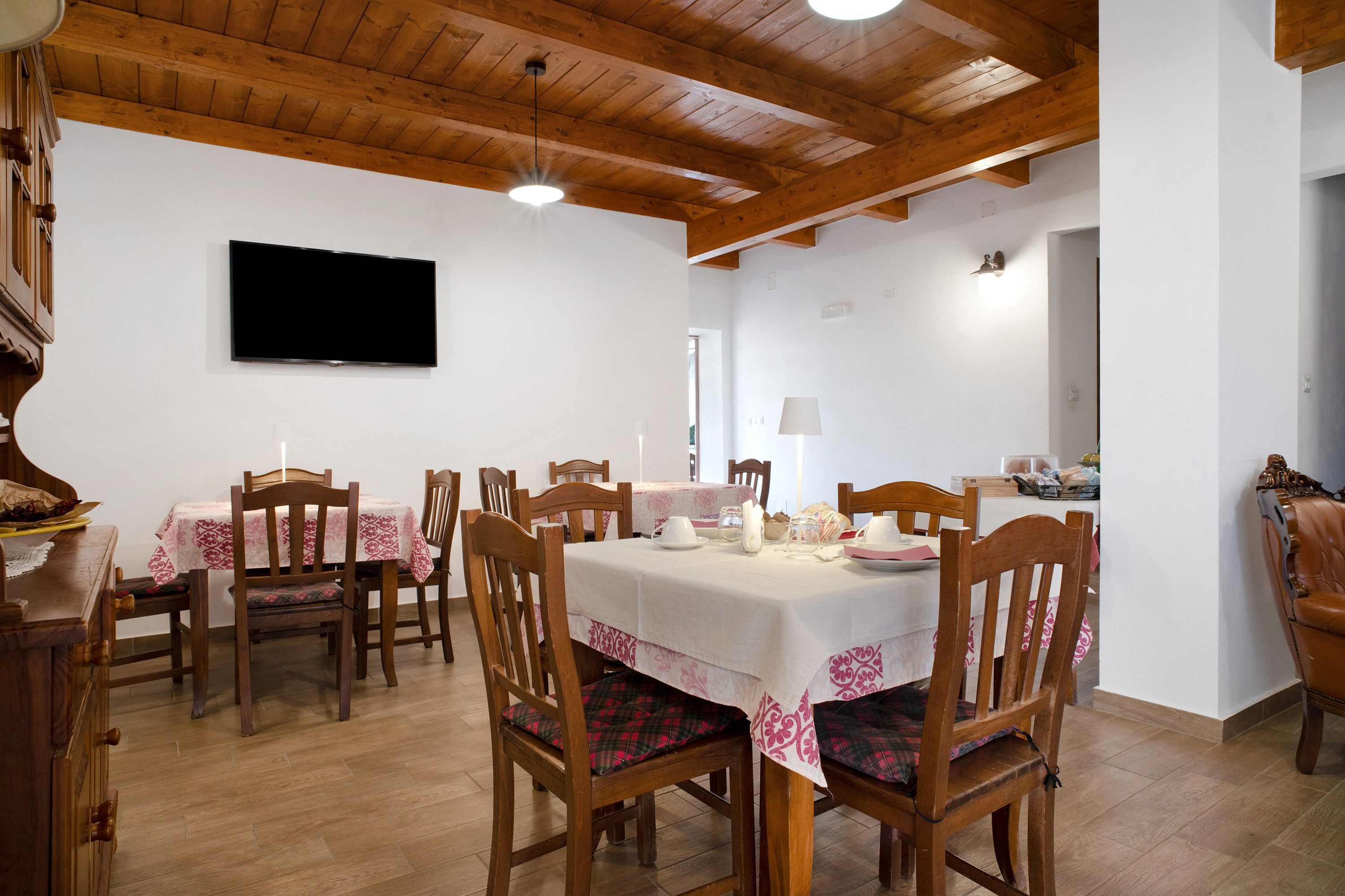 A dining area with wooden tables and chairs, a flat-screen TV mounted on the wall, and a wooden ceiling.