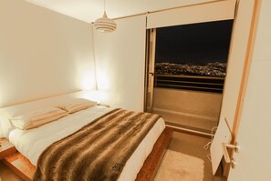 Un dormitori amb un llit, una tauleta de nit, una finestra que ofereix vistes a un paisatge urbà de nit i una làmpada penjant.