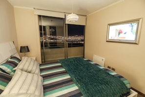 A hotel room with a bed, a view of a cityscape at night, a window with a picture frame, and a lamp. - Viña Lounge (Vina del Mar)