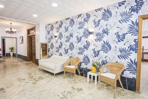 Un hall de hotel con un sofá blanco, dos sillones de mimbre, una mesita con un jarrón y una pared con estampado floral.