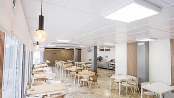 Un grand espace intérieur aménagé en salle à manger, avec de nombreuses tables et chaises, une cuisine à l’arrière-plan et un plafond doté d’un éclairage encastré.