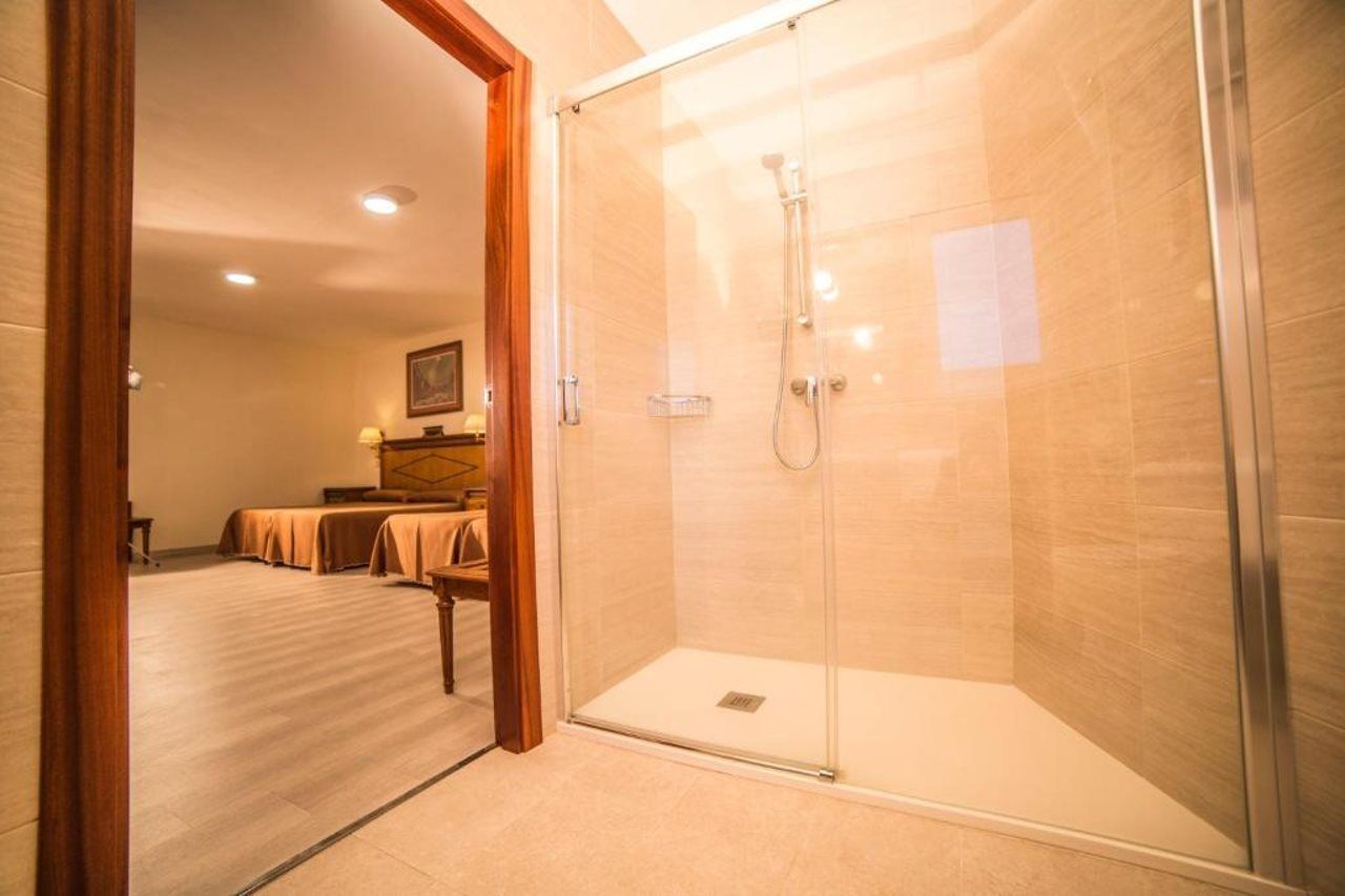 Ein Hotelbad mit einer Glaskabine zur Dusche, einem Bett mit Holz Kopfteil und einem kleinen Tisch mit Lampe.