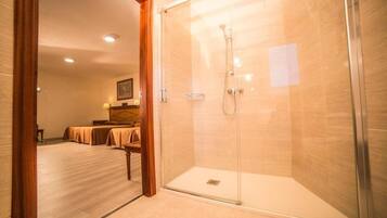 Ein Hotelbad mit einer Glaskabine zur Dusche, einem Bett mit Holz Kopfteil und einem kleinen Tisch mit Lampe.