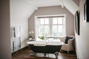 A bathroom with a freestanding bathtub, a window with blinds, a wicker chair, and a radiator. - Woodlands Hotel (Leeds)