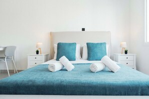 Una cama bien hecha con colcha color turquesa, dos almohadas y toallas blancas enrolladas. Hay dos mesitas de noche con lámparas y pequeñas macetas. Se ve una silla gris a la izquierda.