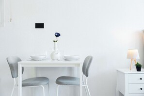 Un comedor minimalista con una mesa blanca, dos sillas grises, un jarrón con una sola flor y una mesita de noche con una lámpara.