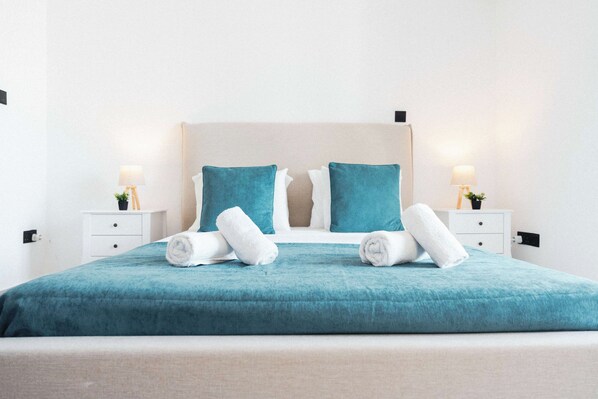 Un lit bien fait, avec une couvre-lit turquoise et des oreillers blancs, deux serviettes blanches roulées et une tête de lit en bois.