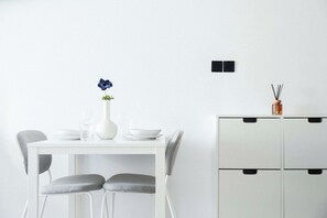A minimalist dining area with a white table, two grey chairs, a vase with a single flower, and a cabinet with black handles. - Modern Central Studio Walk to Beach & Dining (Lemesos)