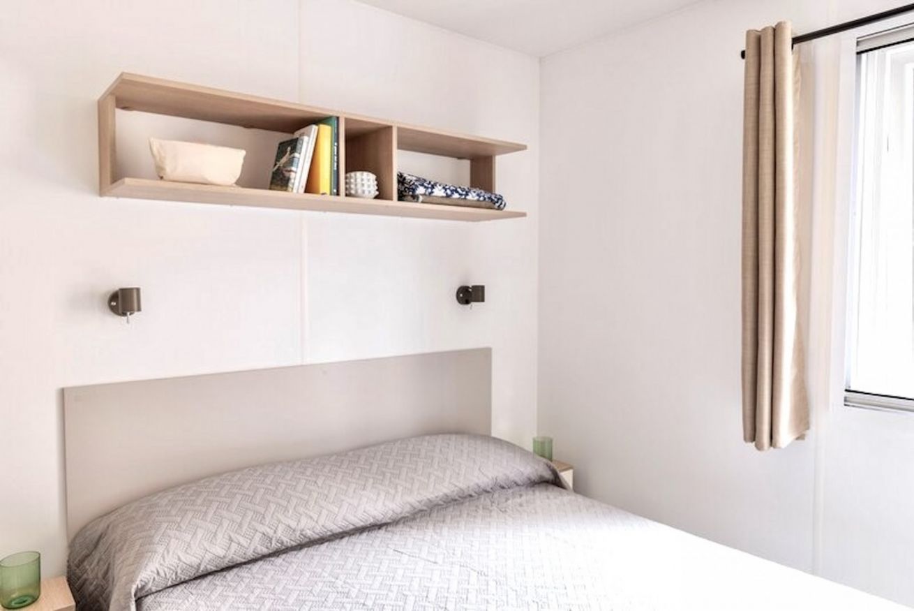 A bedroom with a bed, a shelf above it, and a window with curtains.