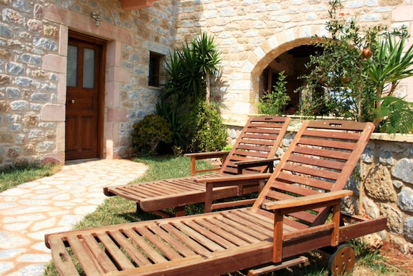 A stone building with a wooden door and a small window. There are two wooden lounge chairs with metal frames and a small table. - Mani Village Villas (West Mani)