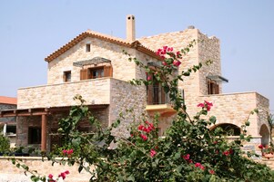 Ένα κτίριο από πέτρα με μπαλκόνι, καμινάδα και ένα παρτέρι με ροζ λουλούδια.
