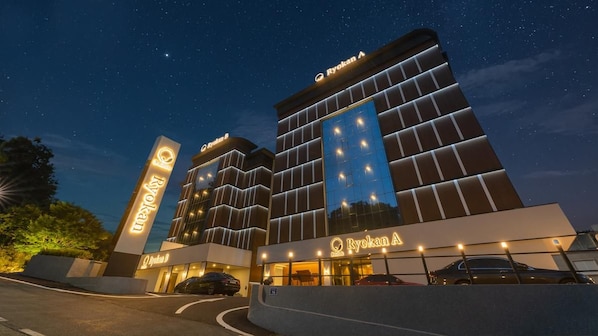 A hotel with a modern facade, illuminated signage, and a parking area. - Denbasta Ryokan Hotel (Geoje)