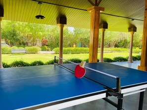 Mesa de ping pong al aire libre con red y una paleta roja, rodeada de pilares de madera y un techo verde.