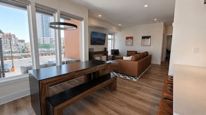A modern living room with a wooden dining table, a sofa, and a view of the city through large windows. - The James Suite Two | New 2BR Beach Block (Atlantic City)