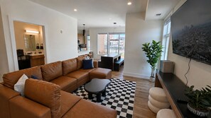 A modern living room with a brown leather sofa, a black and white checkered rug, and a large flat-screen TV mounted on the wall. - The James Suite Two | New 2BR Beach Block (Atlantic City)