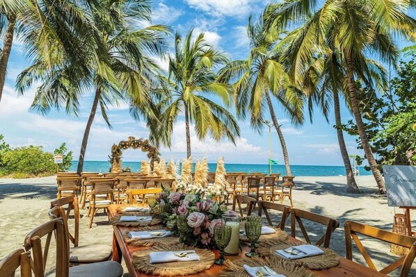 An outdoor dining area with tables set for a meal, surrounded by palm trees and overlooking a beach. - Irotama Resort Zona Torres (Santa Marta)