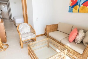 Una habitación con sofá, una silla de mimbre, una mesa de centro de vidrio y una cama al fondo.