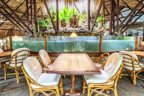 Un restaurante con mesa de madera, sillas de mimbre, techo de paja y una fuente de agua.