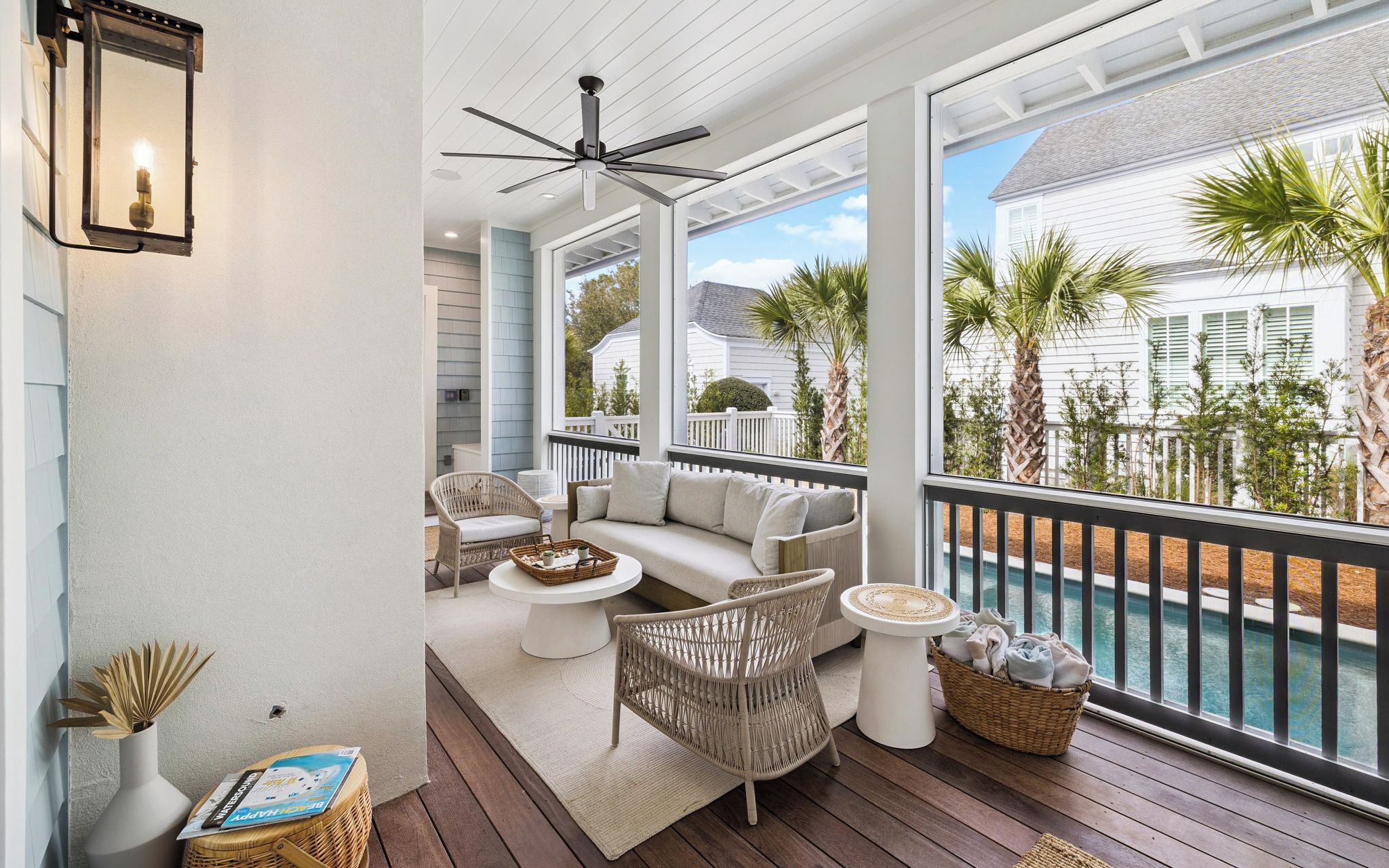 A covered outdoor area with a ceiling fan, wicker furniture, a white sofa, and a view of a pool and white house.