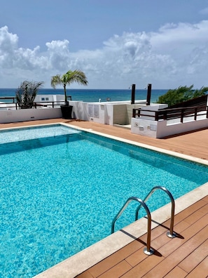 A clear blue pool with a wooden deck, a palm tree, and a view of the ocean. - New Listing! Beachfront 2BR with Ocean View, Pool & Parking (Playa del Carmen)